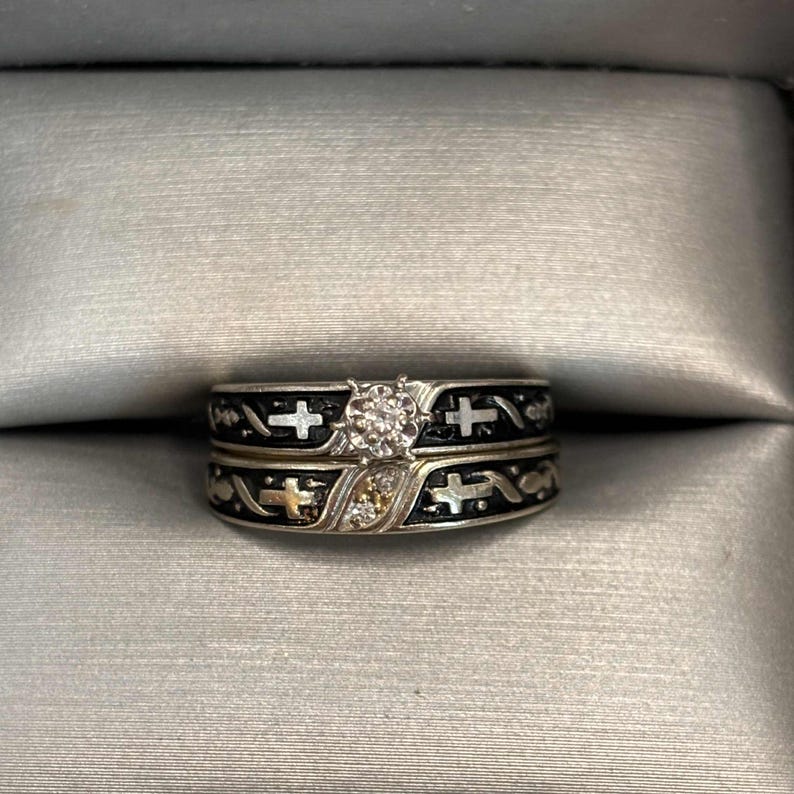 May include: A silver wedding ring set with a diamond centre stone. The bands feature a black enamel design with cross and decorative accents. The rings are displayed in a grey ring box.