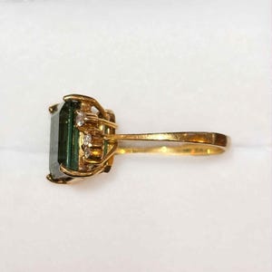 May include: A gold ring featuring a large, rectangular, dark green gemstone. The ring is accented with small, clear gemstones on the sides. The band and setting are gold-colored.