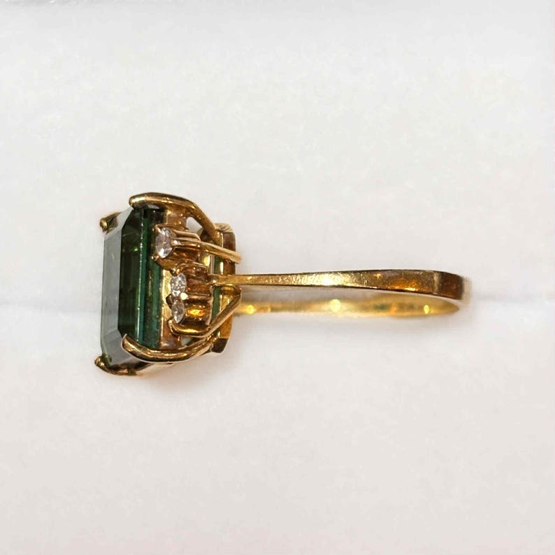 May include: A gold ring featuring a large, rectangular, dark green gemstone. The ring is accented with small, clear gemstones on the sides. The band and setting are gold-colored.