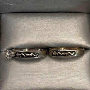 May include: Two silver rings presented in a grey box. One ring has a large, clear gemstone. Both rings feature a black design with a cross and a curved line. These rings are likely for a wedding or engagement.