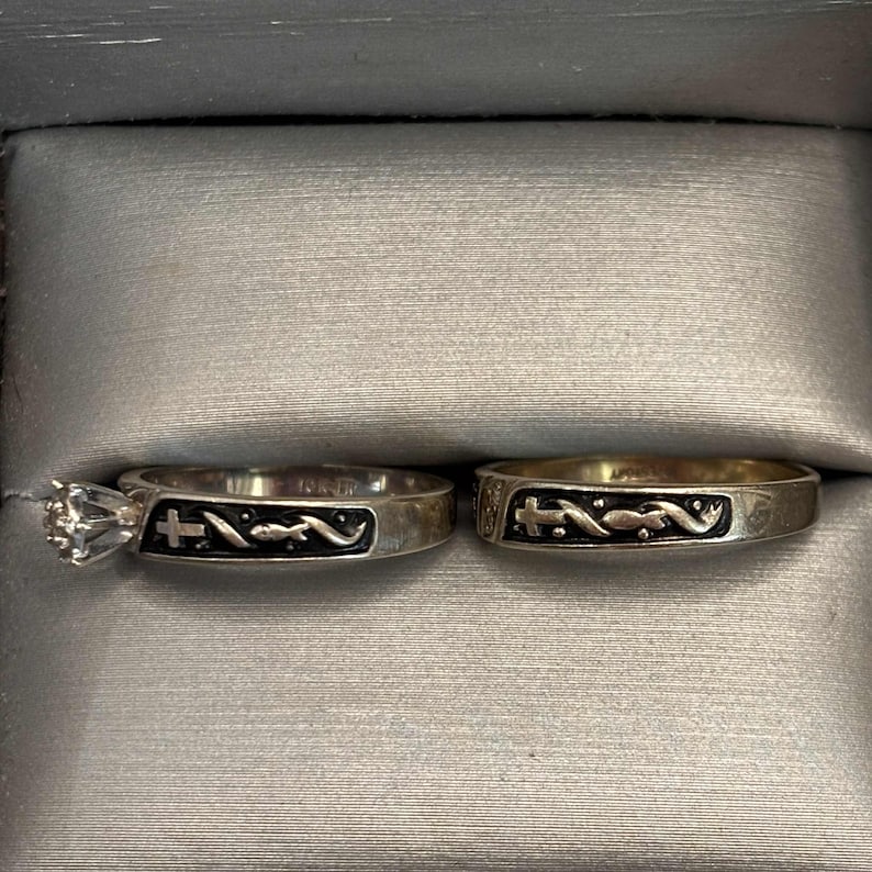 May include: Two silver rings presented in a grey box. One ring has a large, clear gemstone. Both rings feature a black design with a cross and a curved line. These rings are likely for a wedding or engagement.