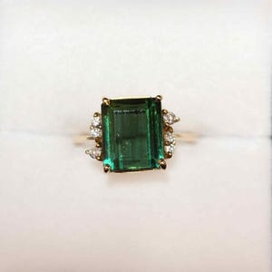May include: A gold ring featuring a large, emerald-cut green gemstone as the centerpiece. The ring is accented with smaller, clear gemstones on either side of the main stone. The setting is gold-colored, and the ring is displayed against a white background.