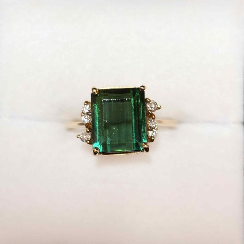 May include: A gold ring featuring a large, emerald-cut green gemstone as the centerpiece. The ring is accented with smaller, clear gemstones on either side of the main stone. The setting is gold-colored, and the ring is displayed against a white background.