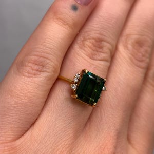 May include: A gold ring featuring a large, emerald-cut, dark green gemstone. The ring is accented with smaller, clear gemstones around the main stone. The ring's band is a thin, polished gold.