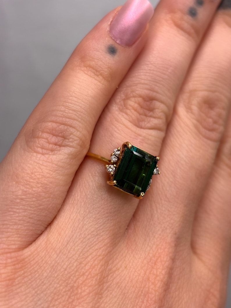 May include: A gold ring featuring a large, emerald-cut, dark green gemstone. The ring is accented with smaller, clear gemstones around the main stone. The ring's band is a thin, polished gold.