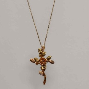 Pode incluir: Um colar dourado com um pendente em forma de cruz com uma rosa no centro e folhas. A cruz &eacute; suspensa por uma corrente fina. O design incorpora elementos florais, criando uma joia elegante e detalhada.