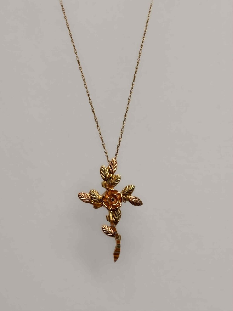 Pode incluir: Um colar dourado com um pendente em forma de cruz com uma rosa no centro e folhas. A cruz &eacute; suspensa por uma corrente fina. O design incorpora elementos florais, criando uma joia elegante e detalhada.