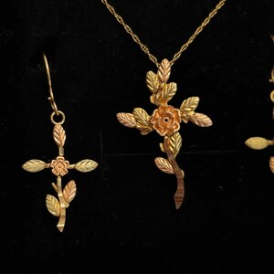 Pode incluir: Um conjunto de joias douradas com um pingente de cruz com um design de rosa e folhas, um par de brincos a condizer e um colar. A cruz e as folhas t&ecirc;m tons de ouro, rosa e verde. As joias est&atilde;o sobre um fundo preto.