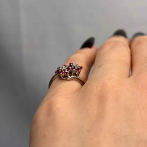 May include: A silver ring featuring a cluster of small, round, red gemstones and clear accent stones. The ring's design is floral-inspired, with the gemstones set closely together. The ring is shown on a hand against a gray background.