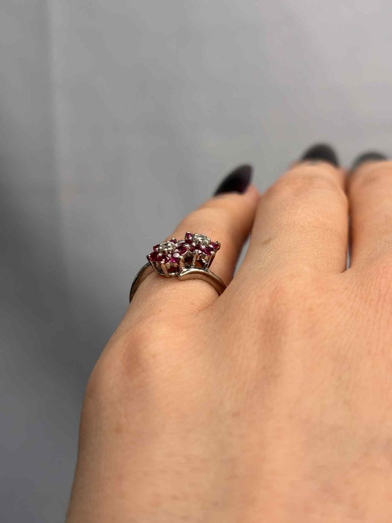 May include: A silver ring featuring a cluster of small, round, red gemstones and clear accent stones. The ring's design is floral-inspired, with the gemstones set closely together. The ring is shown on a hand against a gray background.