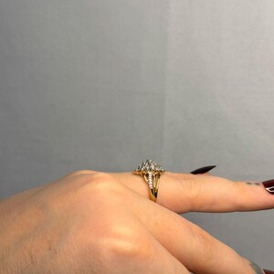May include: A gold ring with a cluster of small, clear stones set in a floral or starburst design. The ring is on a finger, with the band appearing to be split or have a double design. The background is a solid, neutral gray.