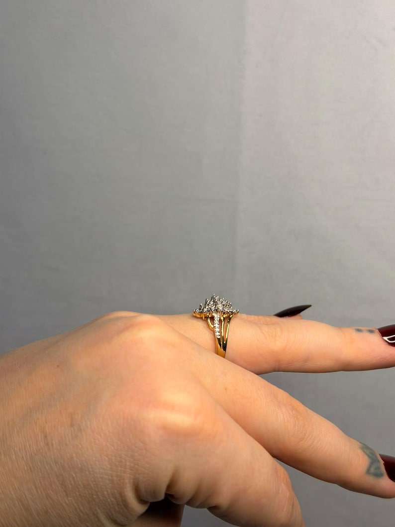 May include: A gold ring with a cluster of small, clear stones set in a floral or starburst design. The ring is on a finger, with the band appearing to be split or have a double design. The background is a solid, neutral gray.