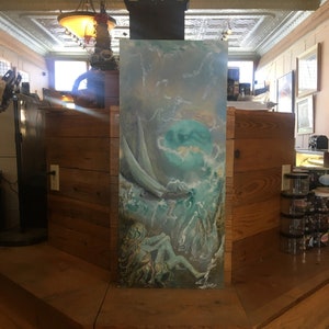 Puede incluir: Una pintura de una sirena con el pelo largo y suelto, sentada en una roca en un mar tormentoso. El mar es de un color azul verdoso vibrante con olas blancas que rompen a su alrededor. La pintura está enmarcada y colgada en una pared.