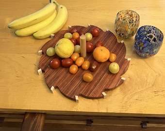 May include: A wooden serving tray with a scalloped edge, featuring a variety of fruit, including bananas, oranges, lemons, and tomatoes. The tray is on a light wood table with two colorful glass cups.