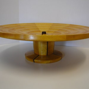 May include: A wooden serving tray with a circular top and a pedestal base. The tray is made of light wood with a natural finish. The base is made of dark wood with a contrasting finish.