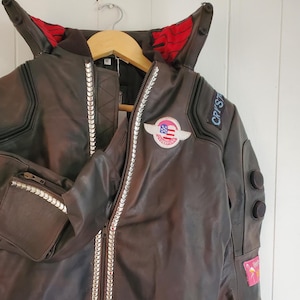 May include: A brown leather jacket with a zipper closure and silver accents. The jacket has a red and black collar with a patch on the left side that says "CRYSTAR" and a patch on the right side with an American flag.
