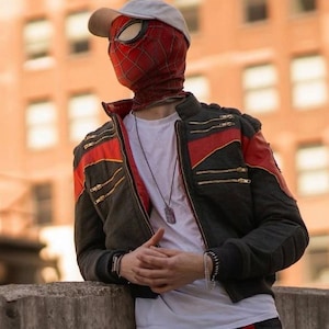 May include: A red and black Spider-Man mask with a white baseball cap, worn with a black jacket featuring red shoulder stripes and gold zippers over a white t-shirt. A silver dog tag necklace and a black beaded bracelet are visible on the chest and wrist.