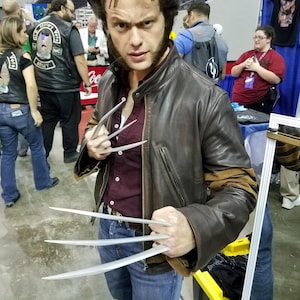 May include: A person dressed as Wolverine, wearing a brown leather jacket, a maroon shirt, and blue jeans. The person is holding three silver claws in each hand, with a stern expression. The hair is styled in the character's signature look.