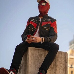 May include: A red and black Spider-Man mask with a web pattern, a beige baseball cap, and a black jacket with red accents and gold zippers. The outfit includes black trousers and red, white, and black sneakers, with a smartphone held in the hands.