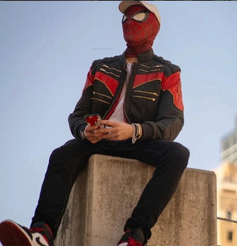 May include: A red and black Spider-Man mask with a web pattern, a beige baseball cap, and a black jacket with red accents and gold zippers. The outfit includes black trousers and red, white, and black sneakers, with a smartphone held in the hands.