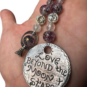 Könnte beinhalten: Eine silberfarbene Halskette mit einem großen, runden Anhänger mit der Aufschrift "Love Beyond the Moon & Stars". Die Halskette hat klare und violette Perlen sowie silberne Charms von Sternen und Monden. Ein kleiner, runder Anhänger mit einem Himmelsdesign ist ebenfalls vorhanden.