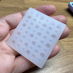 Può includere: Uno stampo quadrato in silicone traslucido con numerose rientranze circolari di varie dimensioni. Lo stampo è tenuto in una mano, mostrando la sua consistenza flessibile e il potenziale per creare piccoli oggetti rotondi.