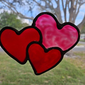 May include: Two overlapping hearts, one red and one pink, with black outlines. The hearts are made of glass and have a stained glass look.