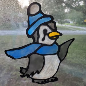 May include: A blue and white penguin wearing a blue hat with a white pom pom and a blue scarf. The penguin is standing on a white surface.