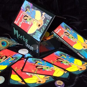 May include: A black box with the words "Meera Tarot" on the front. The box is open and shows a deck of tarot cards with colourful illustrations. The cards are laid out on a black surface with a peacock feather and a white stone.