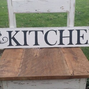 May include: White wooden sign with black lettering that reads "Rowen's Kitchen".