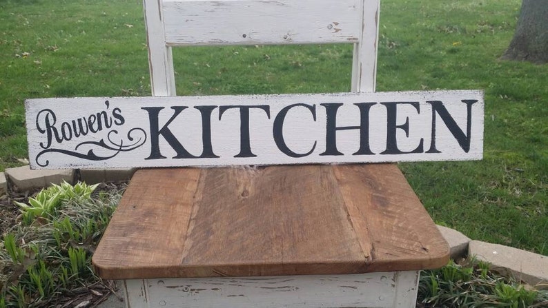 May include: White wooden sign with black lettering that reads "Rowen's Kitchen".