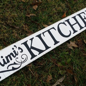 May include: White wooden sign with black lettering that reads "Mimi's Kitchen". The sign is lying on green grass.