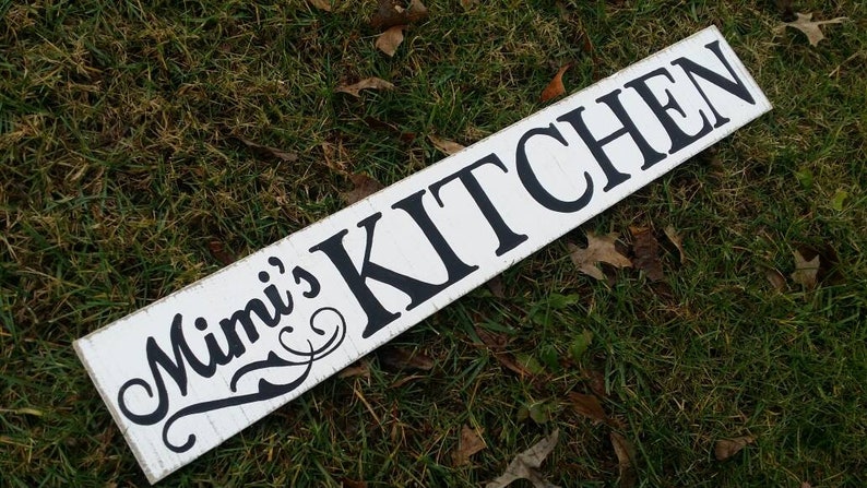 May include: White wooden sign with black lettering that reads "Mimi's Kitchen". The sign is lying on green grass.