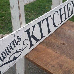 May include: White wooden sign with black lettering that reads "Rowen's Kitchen". The sign is distressed and has a rustic look.