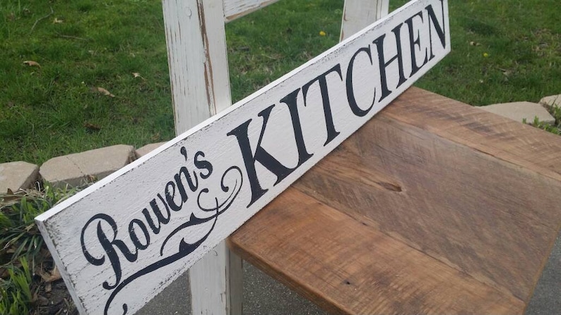 May include: White wooden sign with black lettering that reads "Rowen's Kitchen". The sign is distressed and has a rustic look.