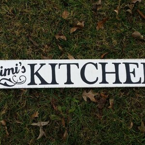May include: White wooden sign with black text that reads "Mimi's Kitchen". The sign is on a bed of green grass.