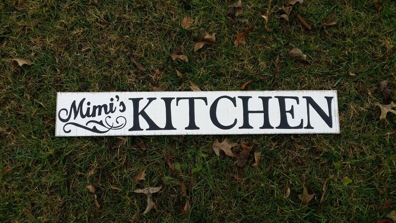 May include: White wooden sign with black text that reads "Mimi's Kitchen". The sign is on a bed of green grass.