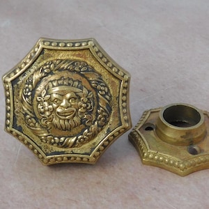 May include: A pair of ornate brass door knobs with a decorative design featuring a face surrounded by foliage. The knobs are octagonal in shape and have a beaded edge.