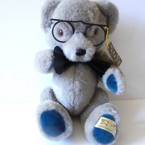 May include: A gray teddy bear wearing black-rimmed glasses and a black bow tie. The bear has blue accents on its paws and a tag attached. The bear is a stuffed animal.
