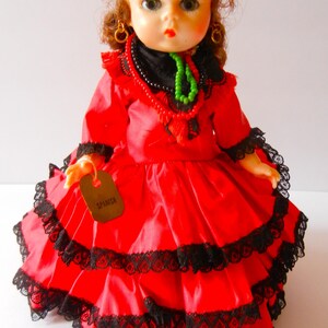 May include: A red and black doll wearing a traditional Spanish dress with a lace trim. The doll has brown hair, green eyes, and is holding a tag that says "Spanish".