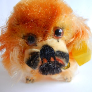 May include: A plush toy dog with orange fur, black eyes, and a black nose. The dog is sitting on a white surface.