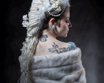 Könnte beinhalten: Eine Person mit weißen Dreadlocks, verziert mit Blumen und Perlen, trägt einen weißen Kunstfell-Schal. Das Bild zeigt Tätowierungen auf Rücken und Schulter, vor dunklem Hintergrund. Die Gesamtästhetik ist künstlerisch und elegant.