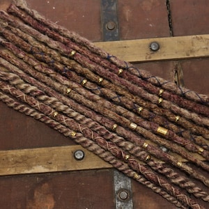 Könnte beinhalten: Eine Ansammlung brauner und burgunderfarbener Dreadlocks mit goldfarbenen Perlen und dekorativen Wicklungen. Die Dreadlocks sind diagonal auf einer dunkelbraunen Oberfläche mit Metallakzenten angeordnet und zeigen einen natürlichen, böhmischen Stil.