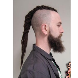 Op de afbeelding: Een man met een geschoren hoofd en een lange, gevlochten dreadlock paardenstaart. Hij heeft een volle baard en draagt een donkergrijs overhemd. Het kapsel is een unieke combinatie van geschoren en dreadlock-elementen.