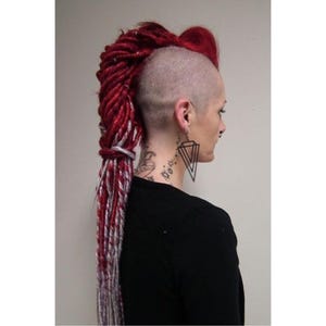 Könnte beinhalten: Eine Person mit rasiertem Kopf und einer auffälligen roten Dreadlock-Frisur. Die Dreadlocks sind zu einem seitlich gefegten Mohawk und Pferdeschwanz mit silbernen Akzenten gestylt. Die Person trägt ein schwarzes Oberteil und geometrische Ohrringe.