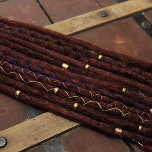 Könnte beinhalten: Burgunderfarbene Dreadlocks mit goldenen Perlen und dekorativen Nähten. Die Dreadlocks sind mit goldfarbenen Perlen und aufwendigen Mustern verziert, was einen einzigartigen und stilvollen Look erzeugt. Die Haarverlängerungen sind dunkelrotbraun.