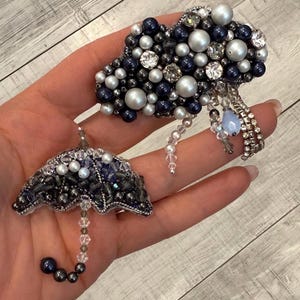 Könnte beinhalten: Handgefertigtes Broschen-Set mit Wolken- und Regenschirm-Design. Die Wolke ist mit marineblauen, silbernen und Perlen verziert, sowie mit funkelnden Kristallen. Der Regenschirm ist mit marineblauen und silbernen Perlen verziert. Ein einzigartiges Accessoire.