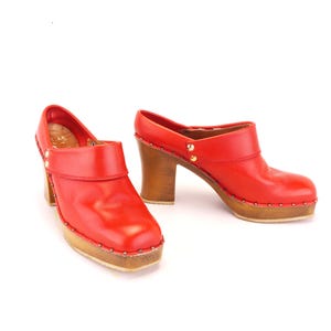 May include: A pair of red leather clogs with wooden heels and soles. The clogs have a rounded toe and are adorned with small gold-colored studs around the edges. The heels are approximately 3 inches high.