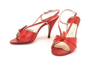 Pode incluir: Um par de sandálias vermelhas de salto alto em couro. Os sapatos de bico aberto têm um detalhe de nó na gáspea e finas tiras no tornozelo. Os saltos têm aproximadamente 7,6 cm de altura.