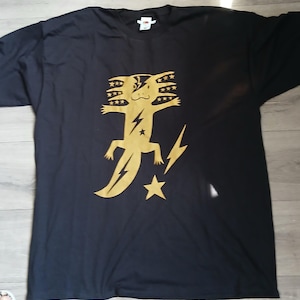Pode incluir: Uma t-shirt preta de manga curta com um grande gráfico dourado de um axolote. O design do axolote inclui um raio sobre um olho, estrelas nas guelras e um símbolo de raio e estrela posicionados à direita da criatura.